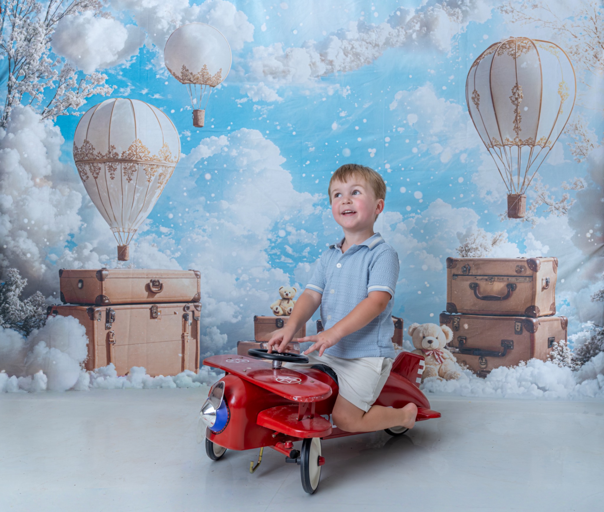 Child sitting on a red toy airplane in front of a sky and hot-air-balloon backdrop