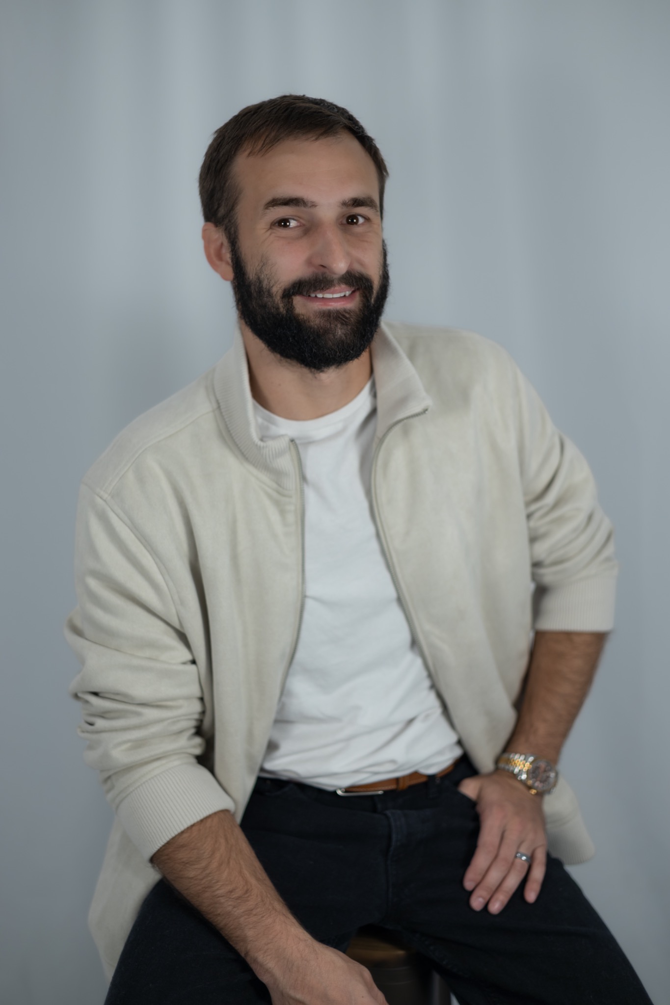 Studio portrait of a smiling bearded man seated in a light overshirt