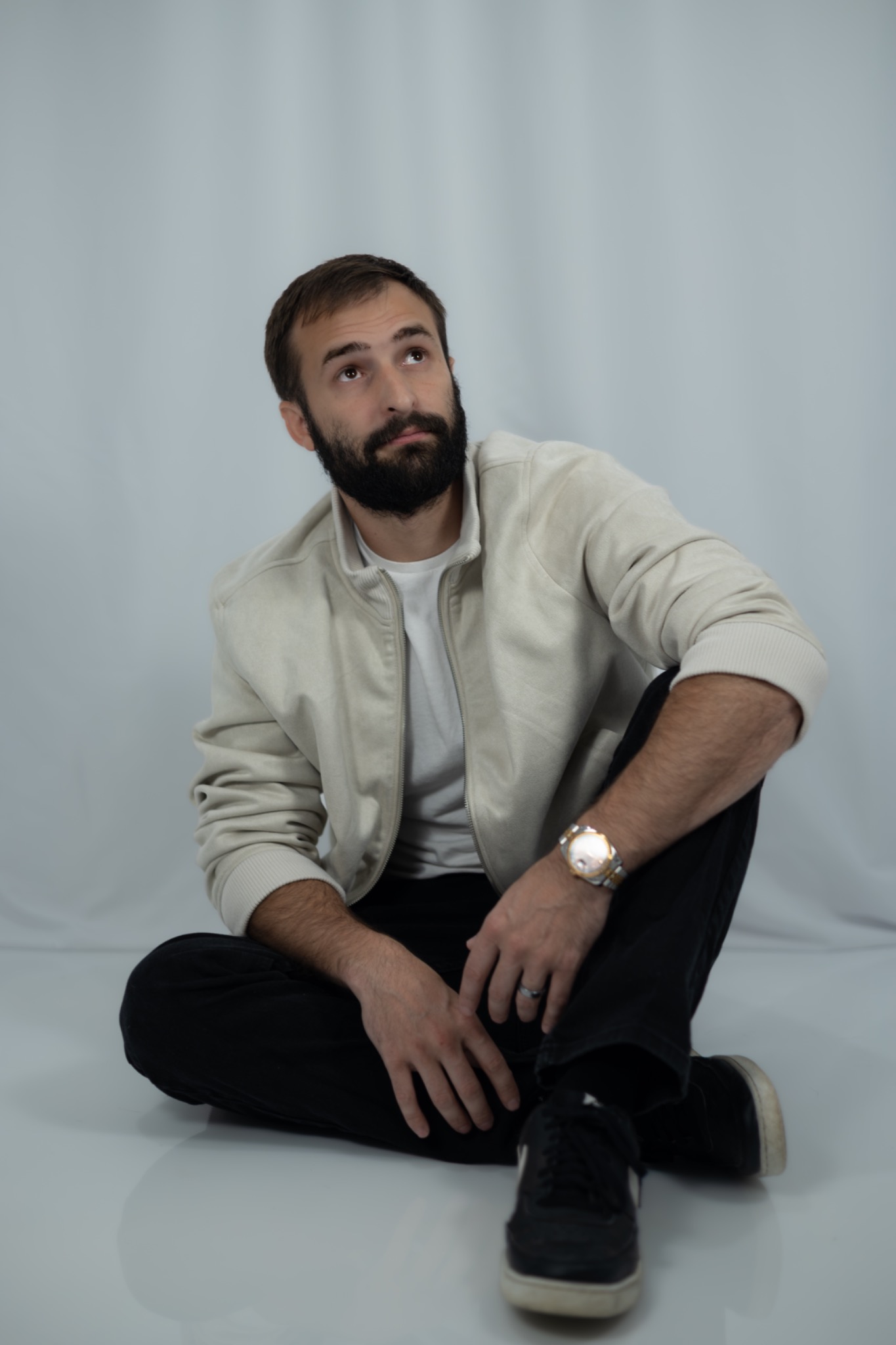 Studio portrait of a bearded man seated on the floor in a light overshirt