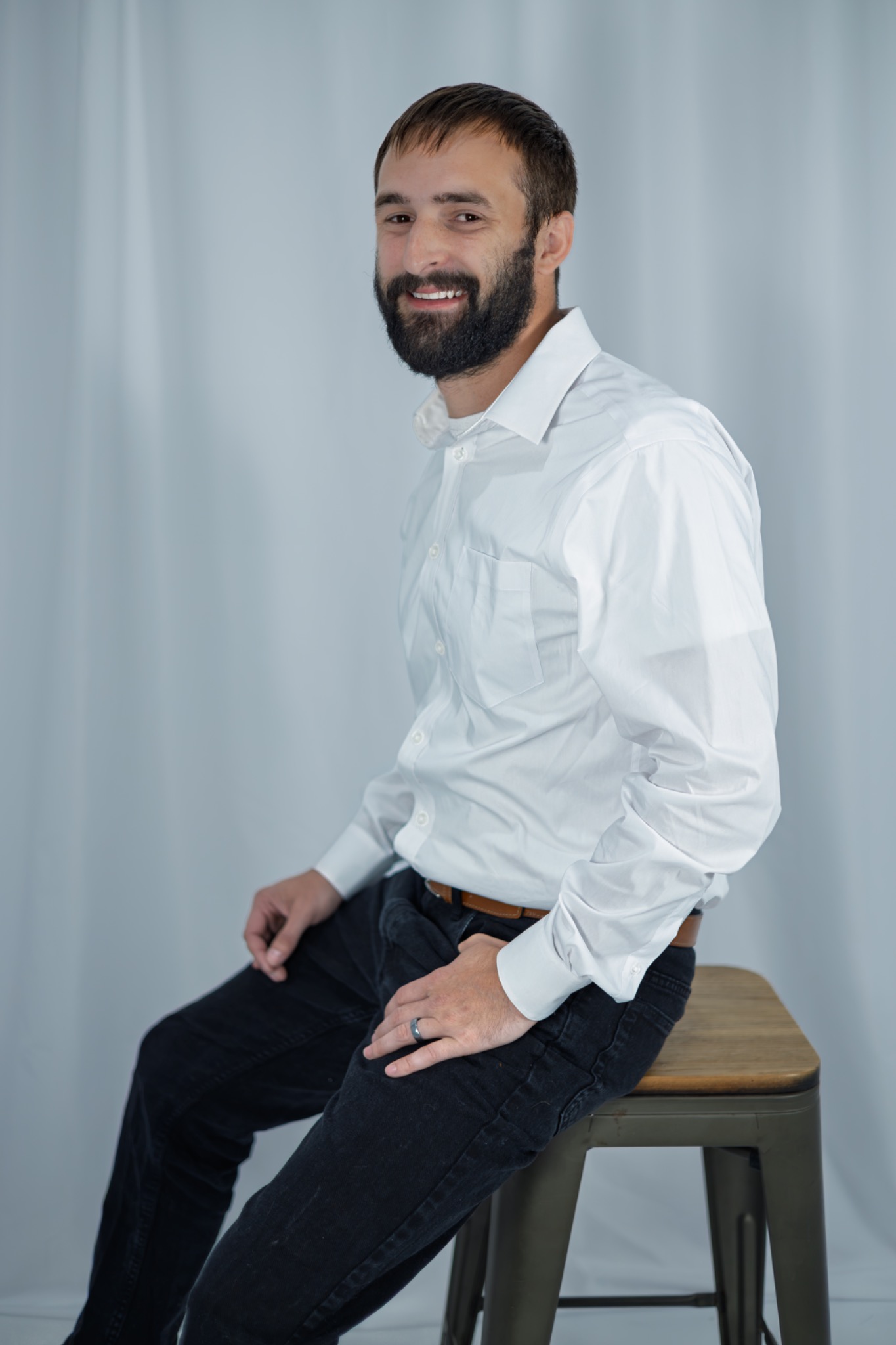Studio portrait of a smiling bearded man seated sideways on a stool in a white shirt