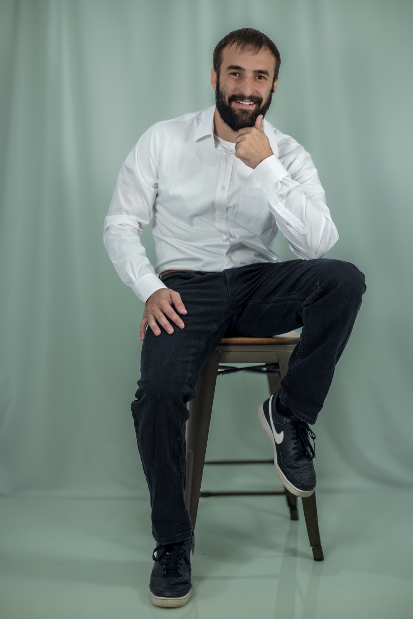 Studio portrait of a bearded man seated on a stool in a white shirt