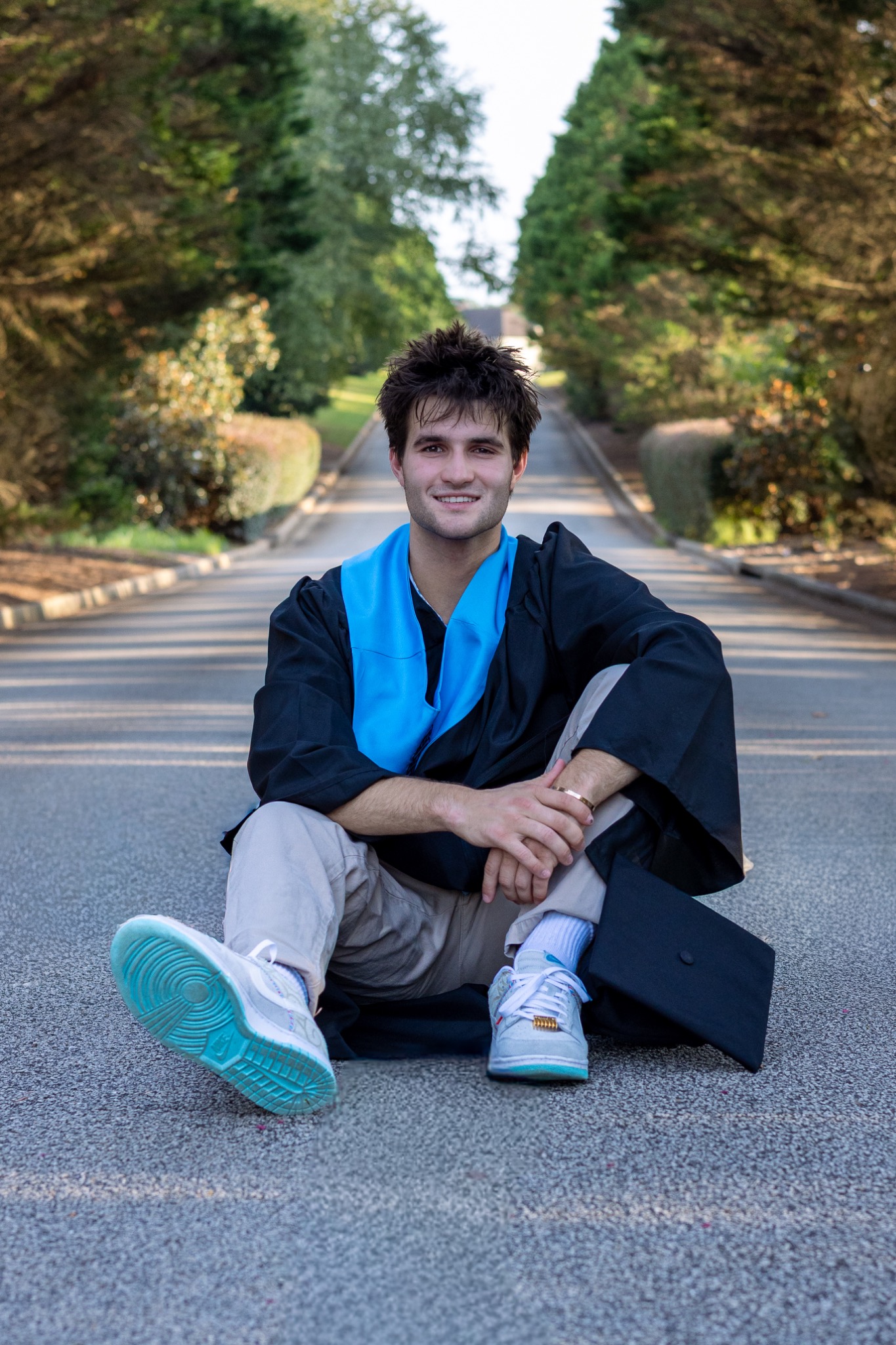 Senior in a graduation gown seated on a tree-lined road