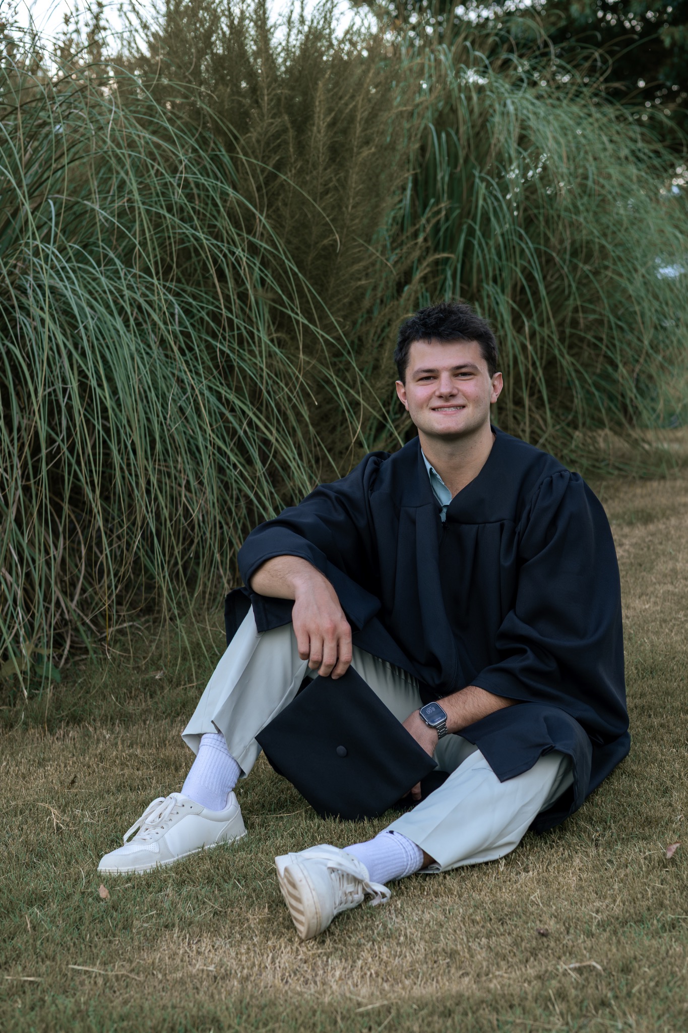 Senior in a graduation gown seated beside tall greenery