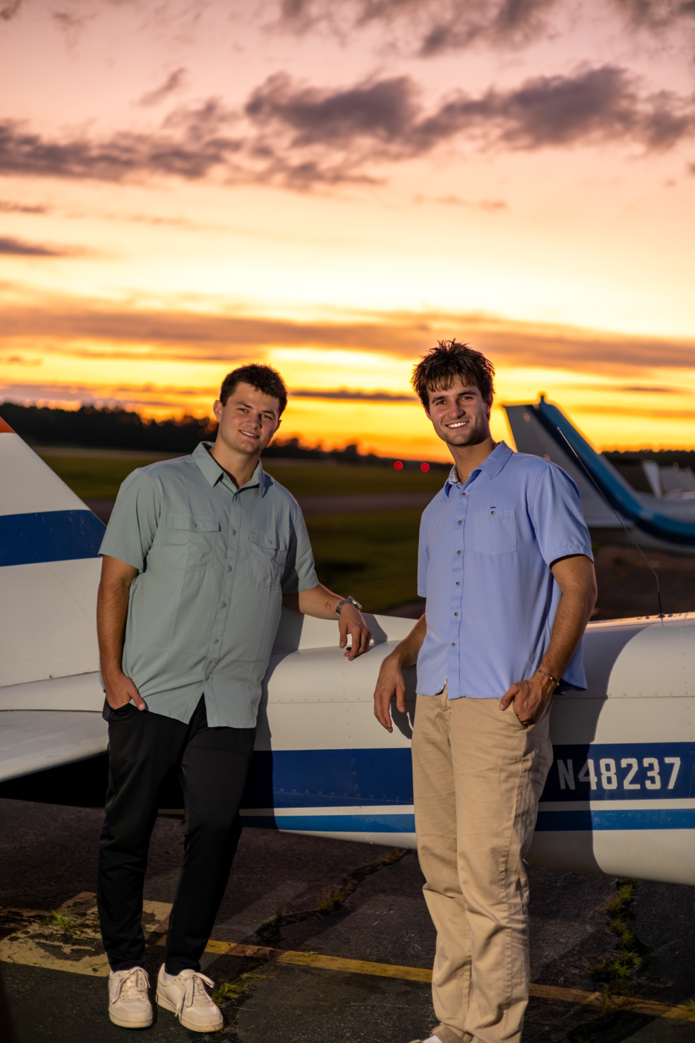Two seniors beside a small airplane beneath a colorful sunset